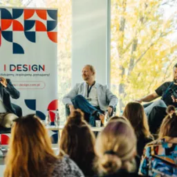 Panel dyskusyjny z czterema prelegentami siedzącymi na fotelach, mikrofony, butelki wody na stole, w tle okno z widokiem na drzewa, widownia z tyłu.