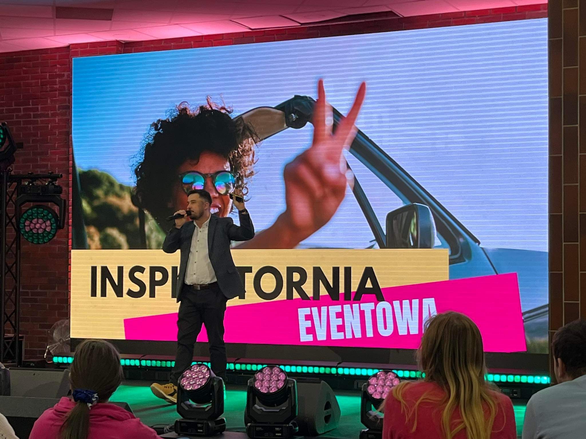 Prezentacja na scenie z mężczyzną w garniturze trzymającym mikrofon, na ekranie za nim wyświetlana jest kobieta w okularach przeciwsłonecznych w kabriolecie pokazująca znak V, obok mężczyzny...