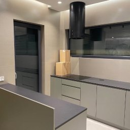 Niebieski Fartuszek - Nowoczesna kuchnia w odcieniach szarości i beżu, z czarnym okapem cylindrycznym i trzema kartonami na blacie, widok na minimalistyczne okno z żaluzjami.