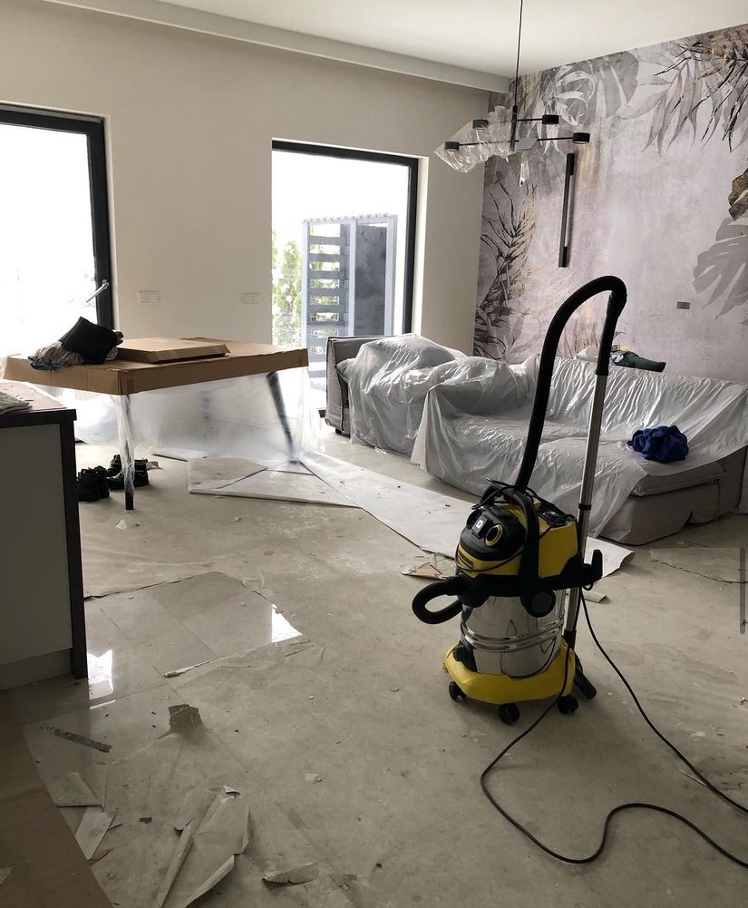 Wnętrze mieszkania w trakcie sprzątania po remoncie, z meblami zabezpieczonymi folią, widoczny żółty odkurzacz przemysłowy i resztki gruzu na podłodze.