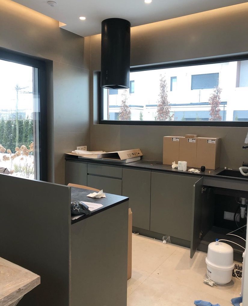 Wnętrze kuchni w trakcie montażu: szare szafki, czarny okap cylindryczny, kartony na blacie, widok na ogród przez okno, biały filtr do wody pod zlewem.