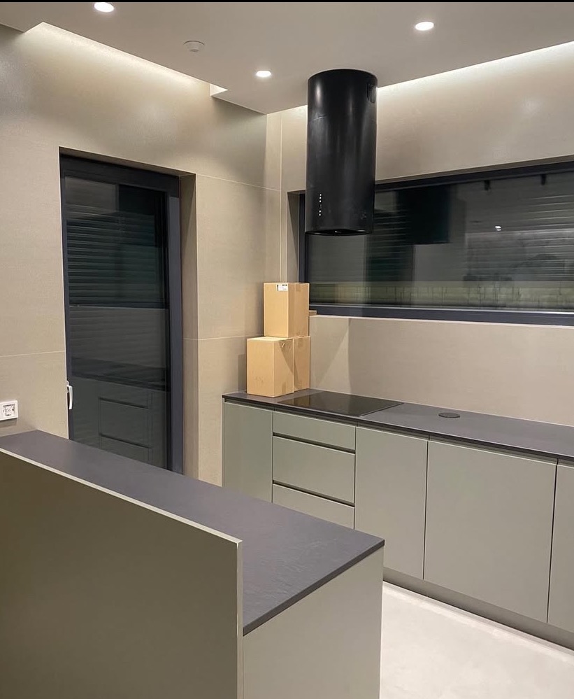 Nowoczesna kuchnia w odcieniach szarości i beżu, z czarnym okapem cylindrycznym i trzema kartonami na blacie, widok na minimalistyczne okno z żaluzjami.