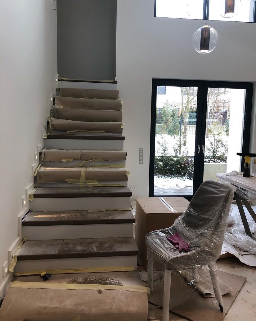 Schody zabezpieczone papierem i taśmą malarską podczas prac remontowych, krzesło owinięte folią ochronną, widoczne ślady pyłu i gruzu.