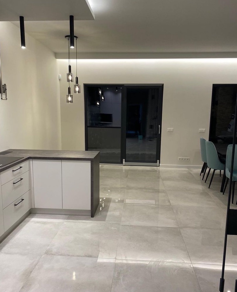 Wnętrze nowoczesnego, minimalistycznego salonu z aneksem kuchennym, jasne płytki podłogowe, białe szafki kuchenne z ciemnym blatem, szklane drzwi przesuwne na taras, wiszące lampy w stylu...