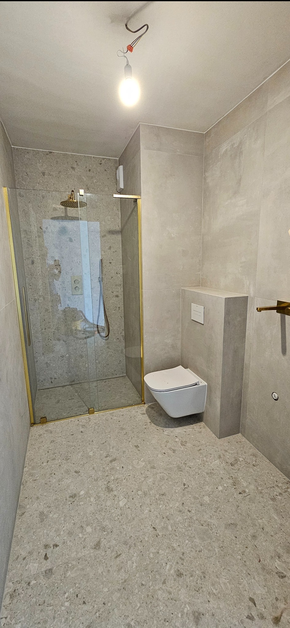 Nowoczesna łazienka z kabiną prysznicową ze złotymi ramami i podwieszaną toaletą, wykończona płytkami w odcieniach szarości z widoczną instalacją elektryczną na suficie.