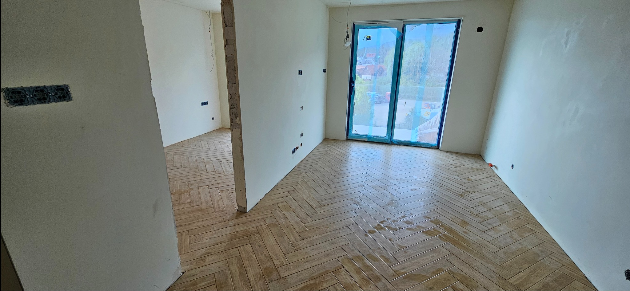 Wnętrze w trakcie wykańczania: świeżo położona podłoga z płytek imitujących drewno w jodełkę, ściany przygotowane do malowania, widoczne puszki elektryczne. Okno balkonowe zabezpieczone folią.