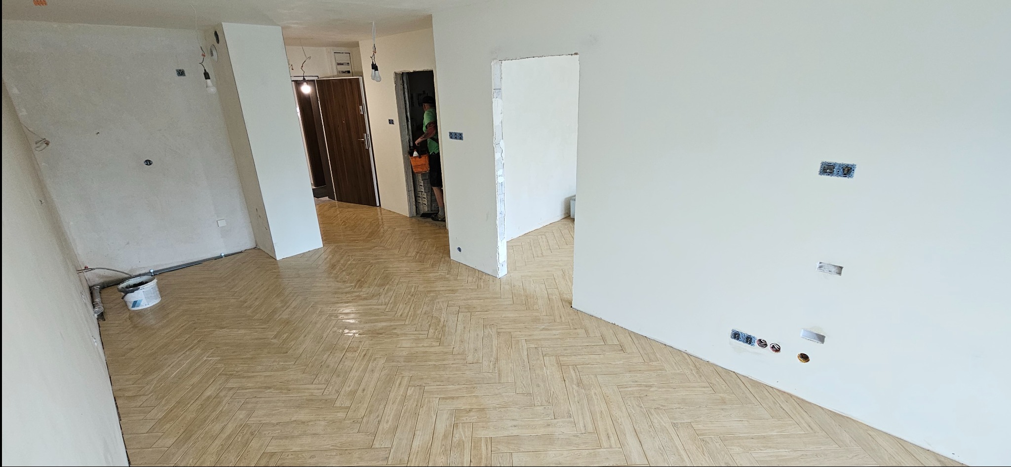 Wnętrze w trakcie prac wykończeniowych: świeżo położona parkietowa podłoga w jodełkę, gładkie, białe ściany z widocznymi puszkami elektrycznymi i otworami po gniazdkach, wiadro z farbą przy ścianie...