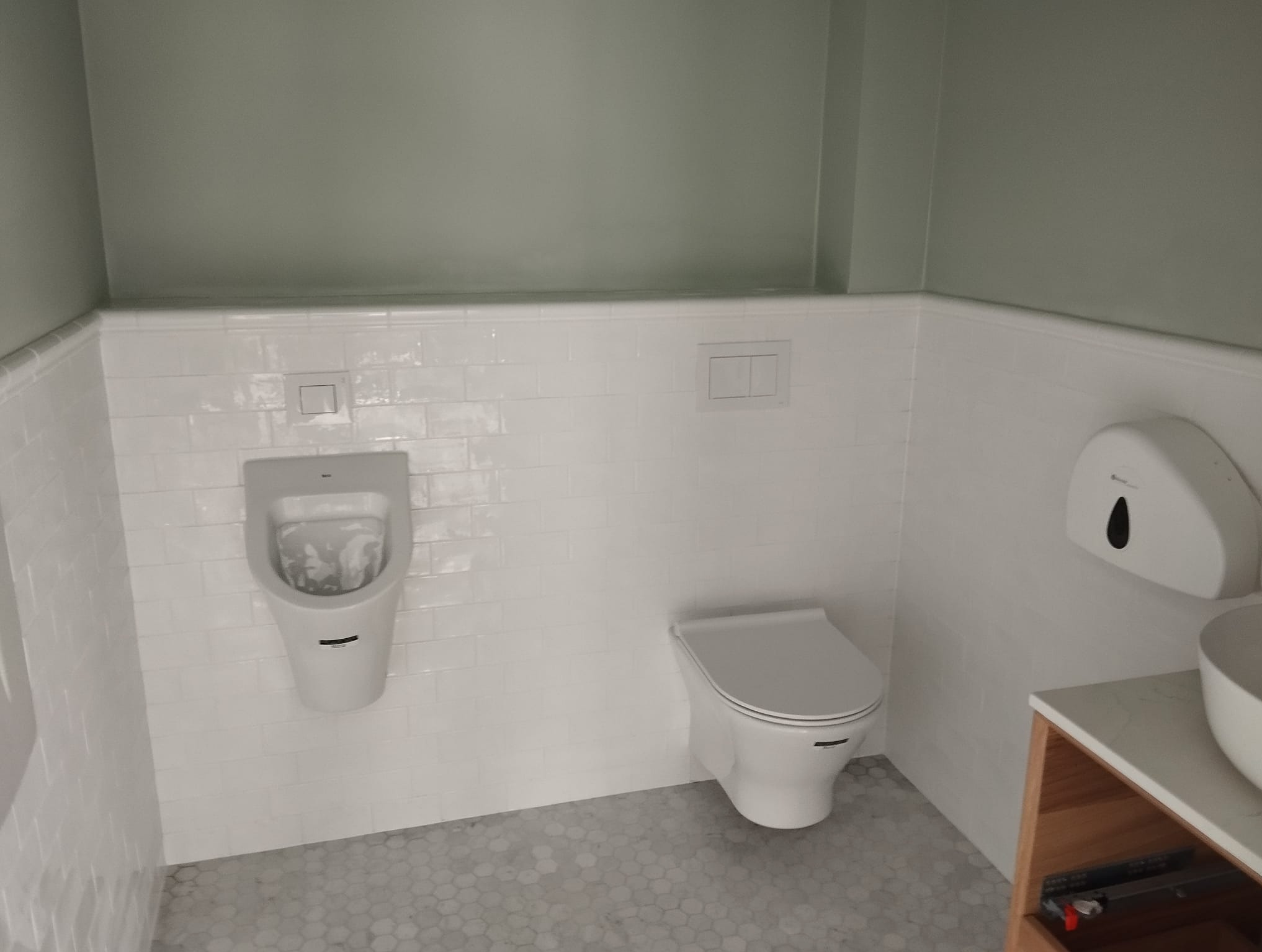 Nowoczesna łazienka z białymi płytkami, wiszącą toaletą, pisuaru i umywalką nablatową na drewnianej szafce, szare heksagonalne płytki podłogowe, minimalistyczny design.