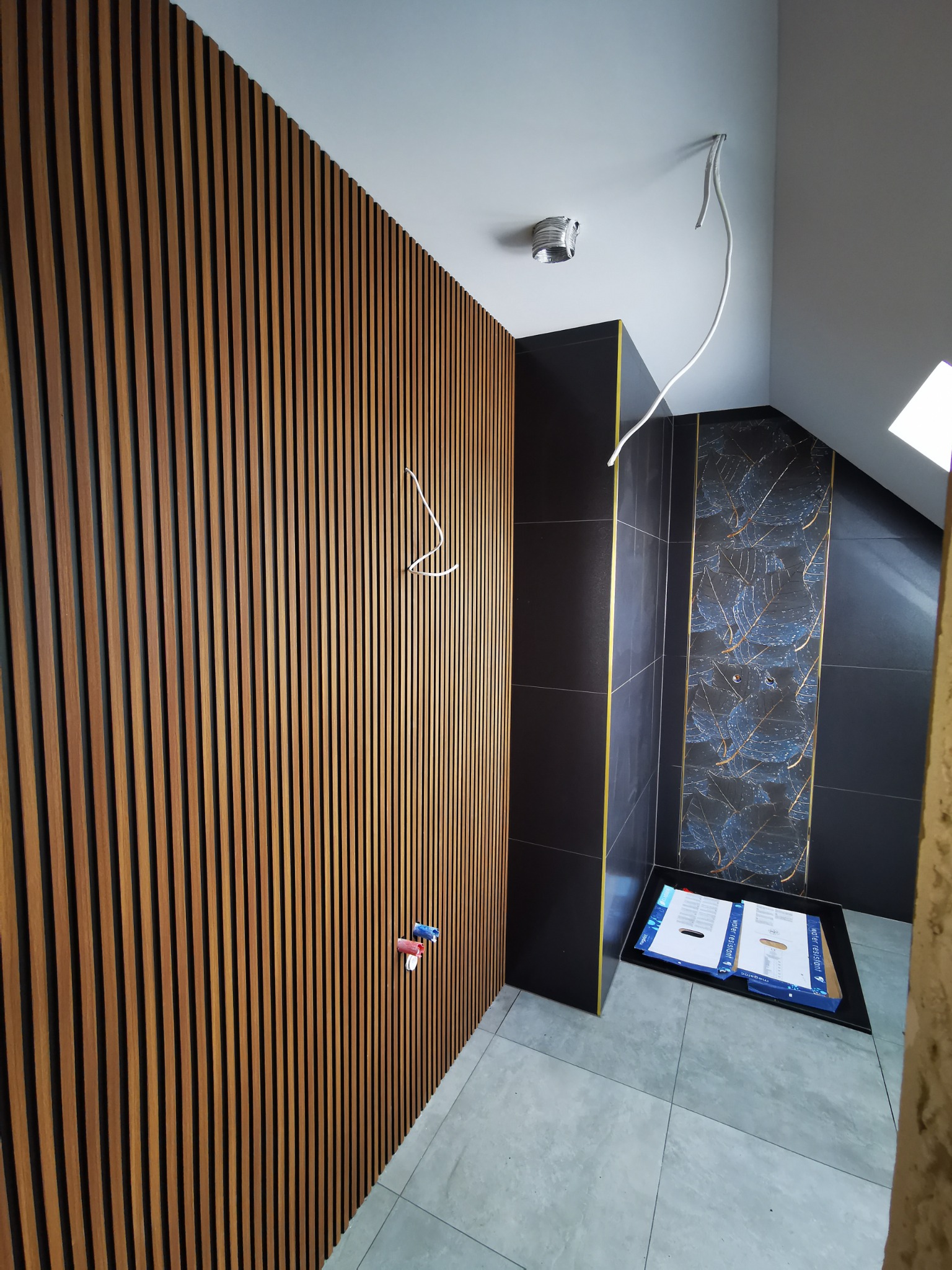 Nowoczesna łazienka w trakcie wykończenia: drewniane lamele na ścianie, czarne płytki z motywem liści, wystające kable i pudełka po płytkach na podłodze.