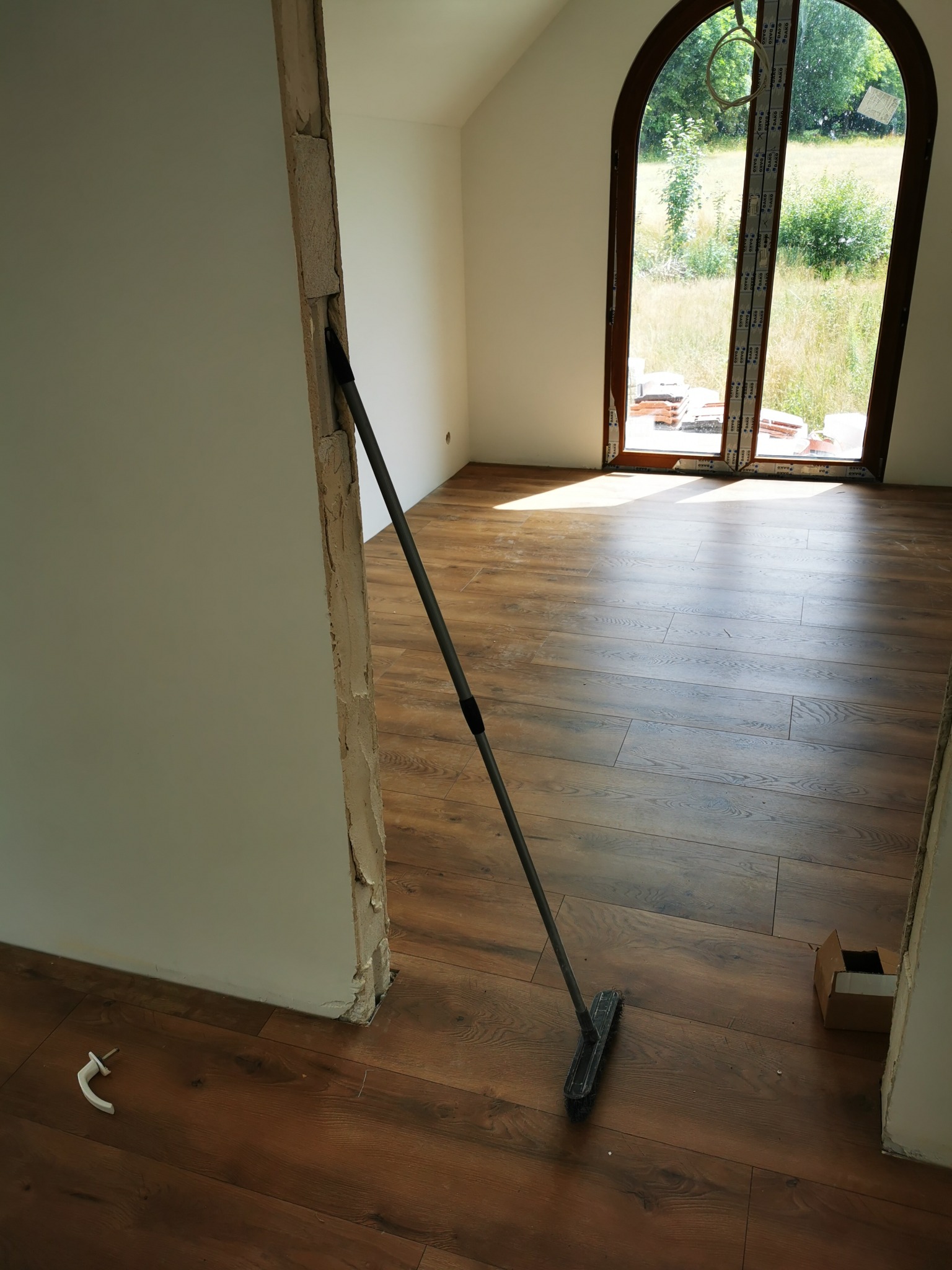 Pokój w trakcie wykańczania, z nową podłogą z paneli, widoczne niedokończone obramowanie drzwi z resztkami zaprawy, okno łukowe z widokiem na zewnątrz oraz szczotka oparta o ścianę, sugerująca...