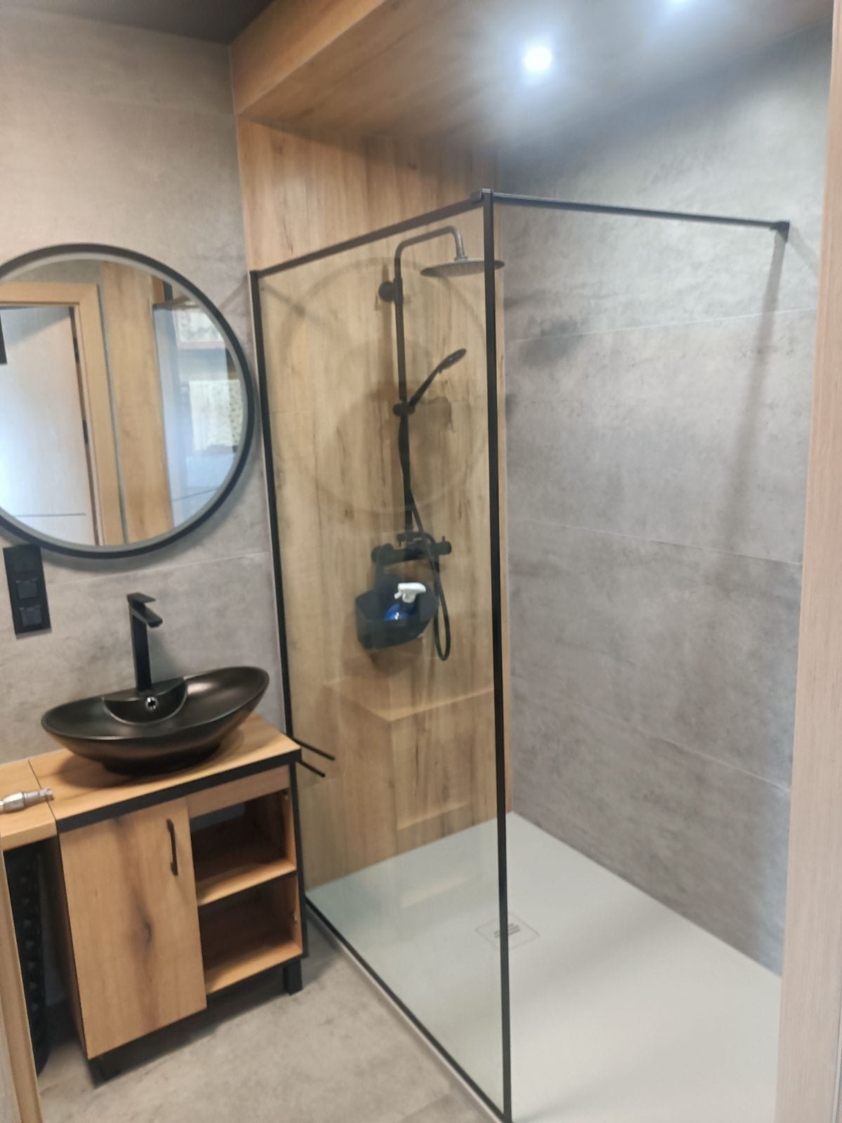 Nowoczesna łazienka z kabiną prysznicową typu walk-in z czarną armaturą, okrągłym lustrem i umywalką nablatową na drewnianej szafce.