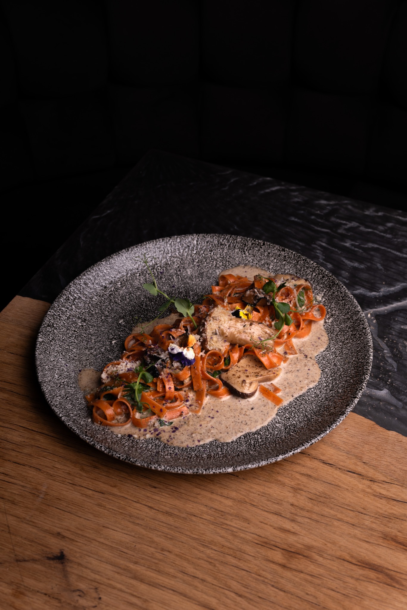 Elegancko podane tagliatelle z sosem grzybowym, posypane serem i ozdobione jadalnymi kwiatami na talerzu o szarej fakturze, na tle ciemnego marmuru i drewnianego blatu.