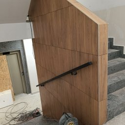 Prime works - Wykończenie klatki schodowej płytkami drewnopodobnymi na ścianie i schodach z szarego lastryko, czarna poręcz, zwinięty przedłużacz na schodach, widoczne kable elektryczne.