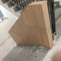 Prime works - Wykończenie klatki schodowej panelami imitującymi drewno, widoczne szare schody z lastryko i czarna metalowa balustrada, w tle okno i biała ściana, na schodach widoczne elementy montażowe.