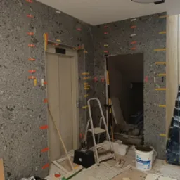 Montaż szarej mozaiki na ścianie przy windzie i schodach, z widocznymi plastikowymi krzyżykami dystansowymi, drabina, wiadro z masą szpachlową i narzędzia glazurnicze na podłodze.