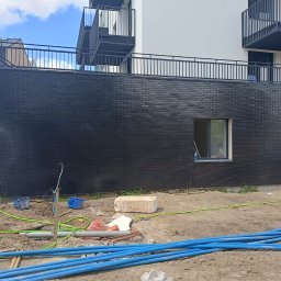 Prime works - Czyszczenie elewacji budynku z czarnej cegły klinkierowej po zakończeniu prac, z widocznymi oknami i balkonami, oraz narzędziami i materiałami budowlanymi na ziemi.