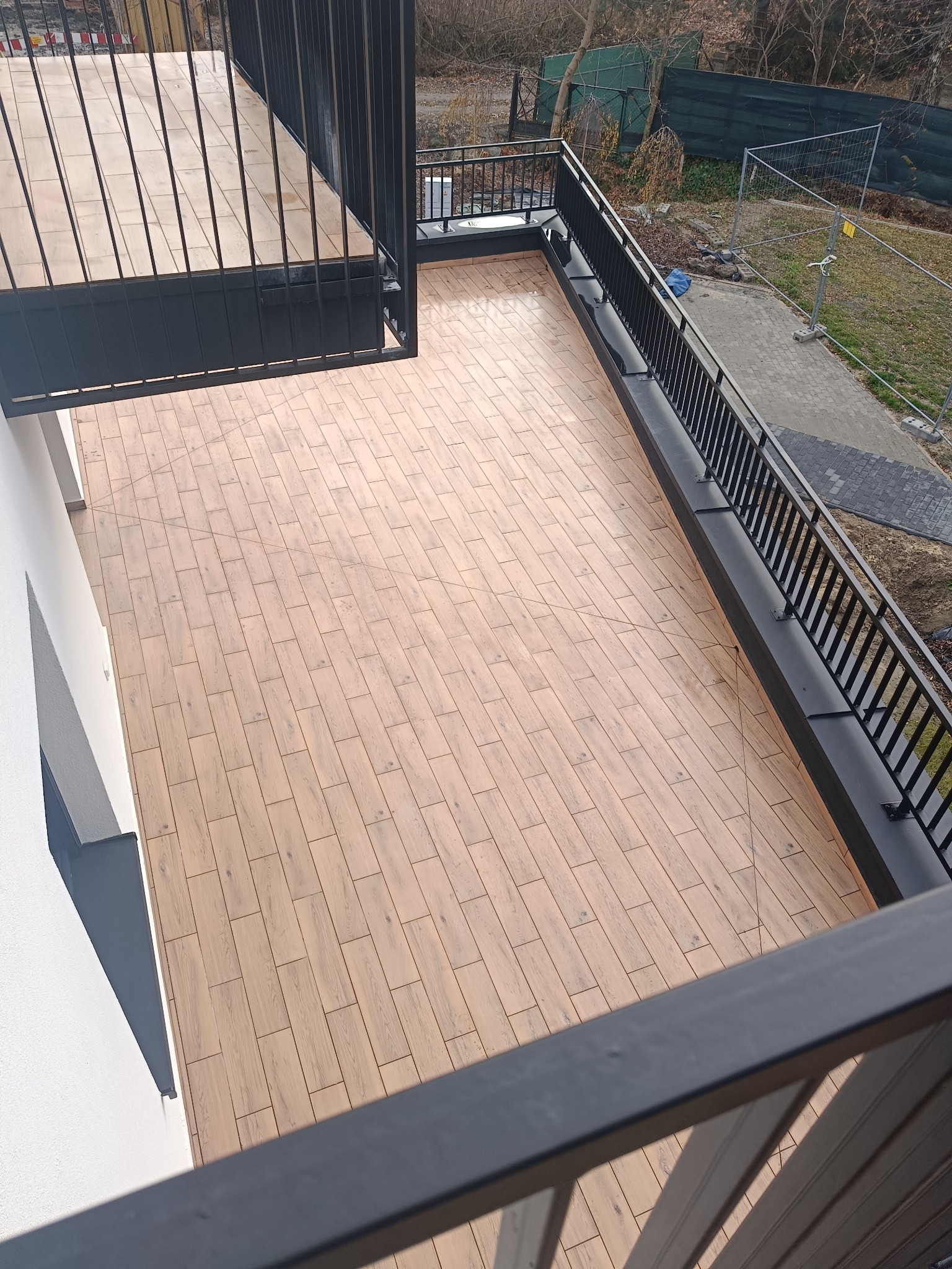 Jasne płytki imitujące drewno ułożone na balkonie z czarną balustradą, widok z góry na ukończoną powierzchnię.