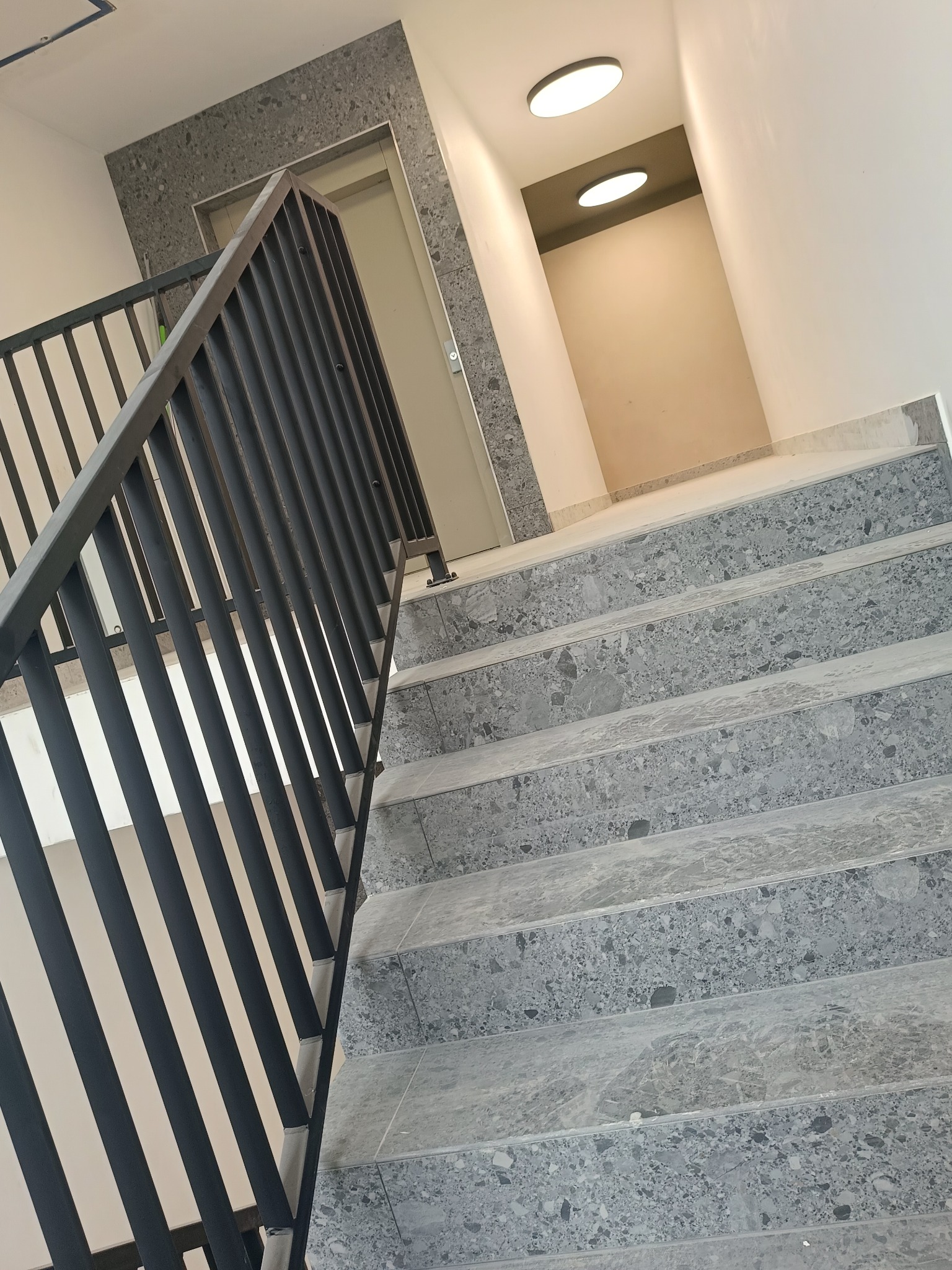 Schody wyłożone szarym lastryko prowadzące w górę, z czarną metalową balustradą, widoczne lampy sufitowe i obramowanie drzwi z lastryko.