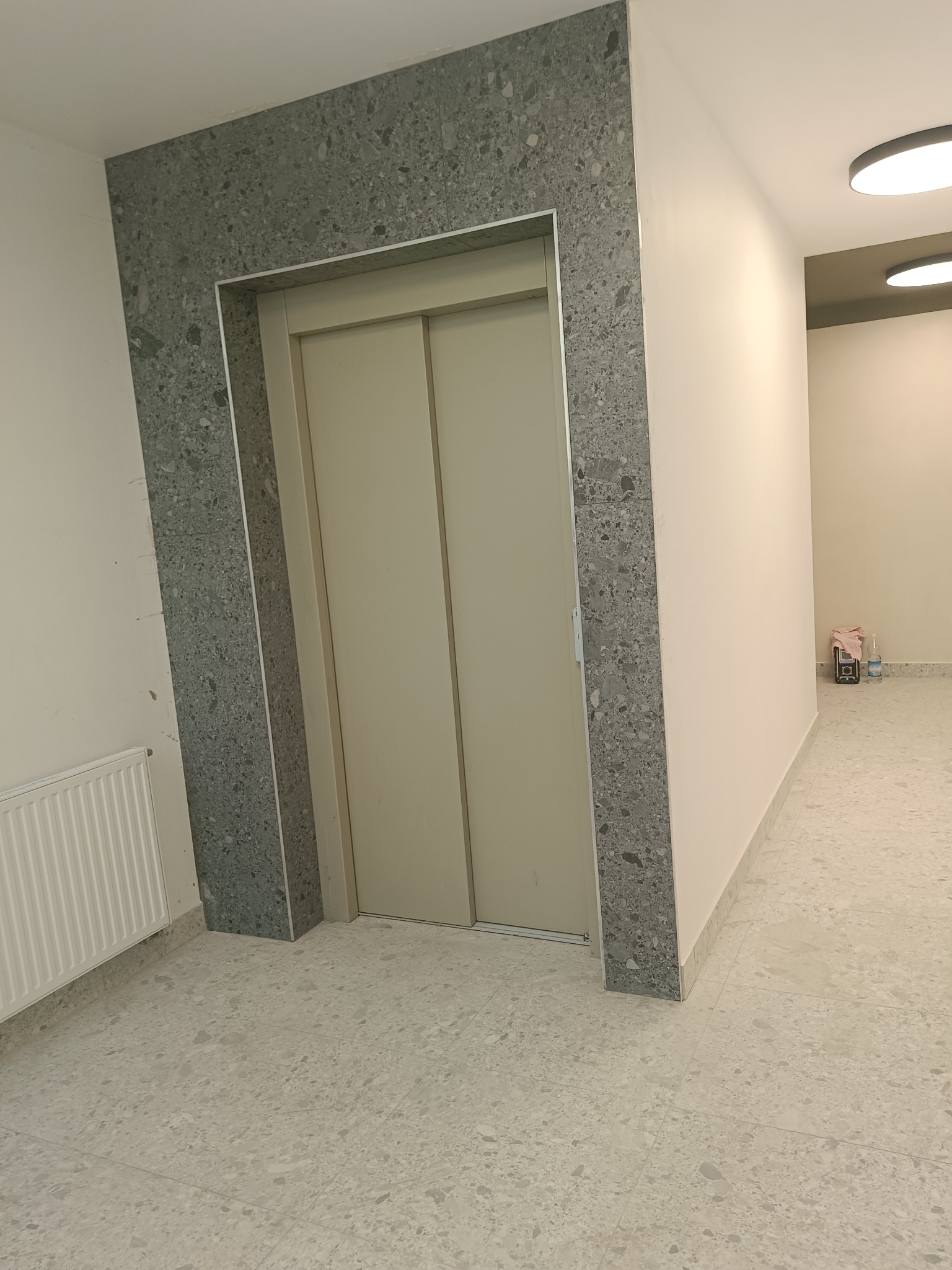 Wykończone wejście do windy z obramowaniem z płytek imitujących lastryko, dopasowanych do posadzki, w jasnym korytarzu.