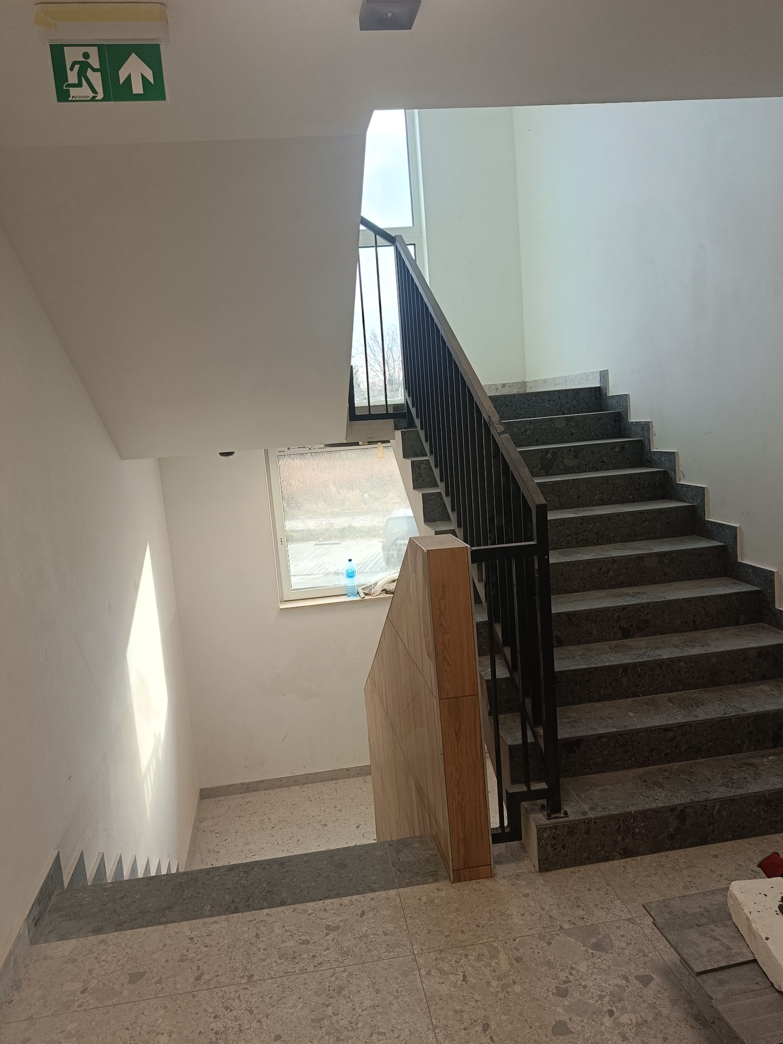 Klatka schodowa w budynku, wykończona szarym gresem, z czarną metalową balustradą. Widoczny znak ewakuacyjny nad schodami i okno z widokiem na zewnątrz. Częściowo widoczne zabezpieczenie schodów...