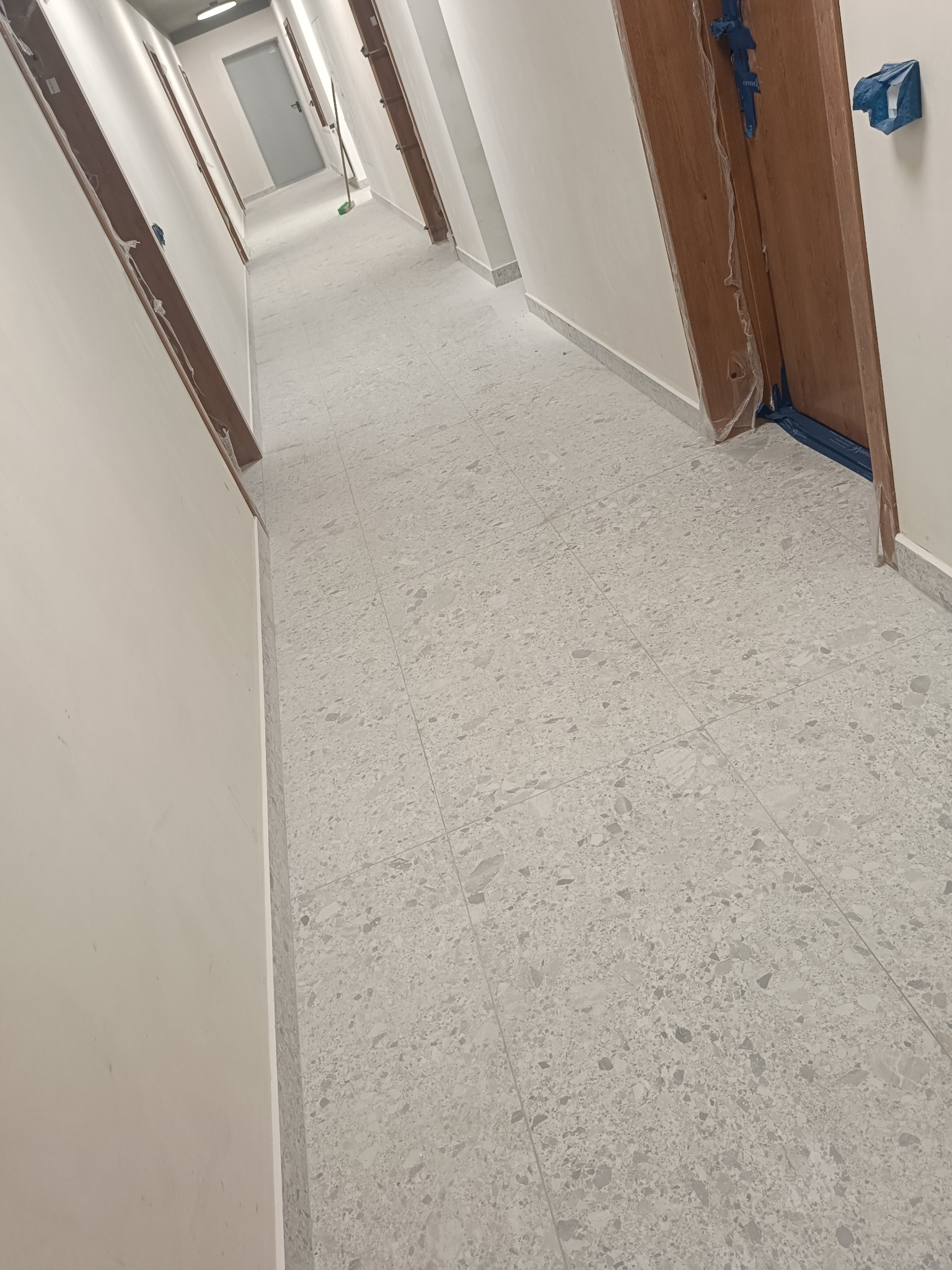 Korytarz w trakcie remontu z położoną jasnoszarą, wzorzystą terakotą na podłodze, zabezpieczonymi folią i taśmą drzwiami oraz przygotowanymi ścianami do malowania.