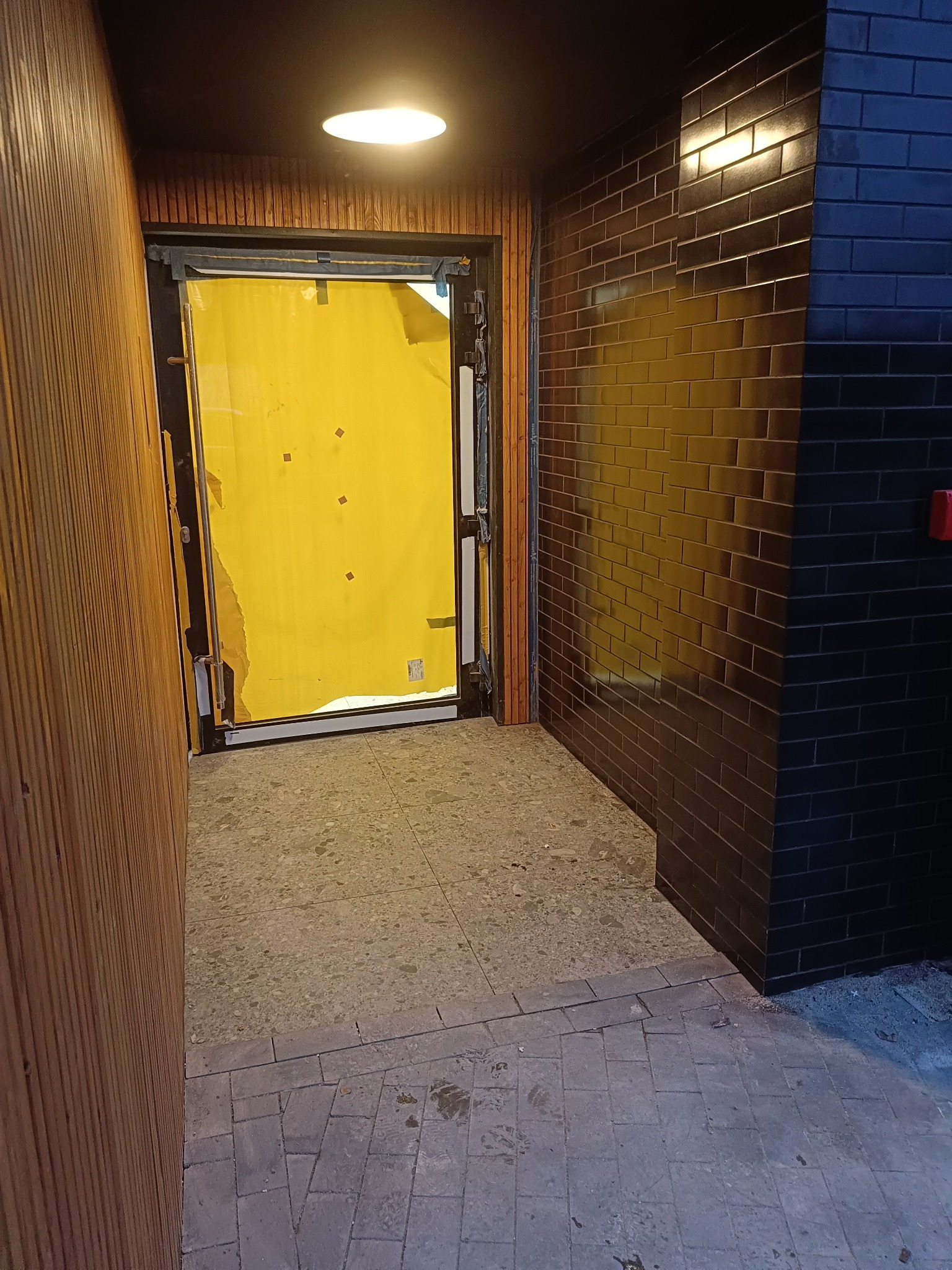 Wejście do budynku w trakcie remontu z żółtą folią zabezpieczającą drzwi, obok ściana wyłożona ciemnymi, błyszczącymi cegłami.