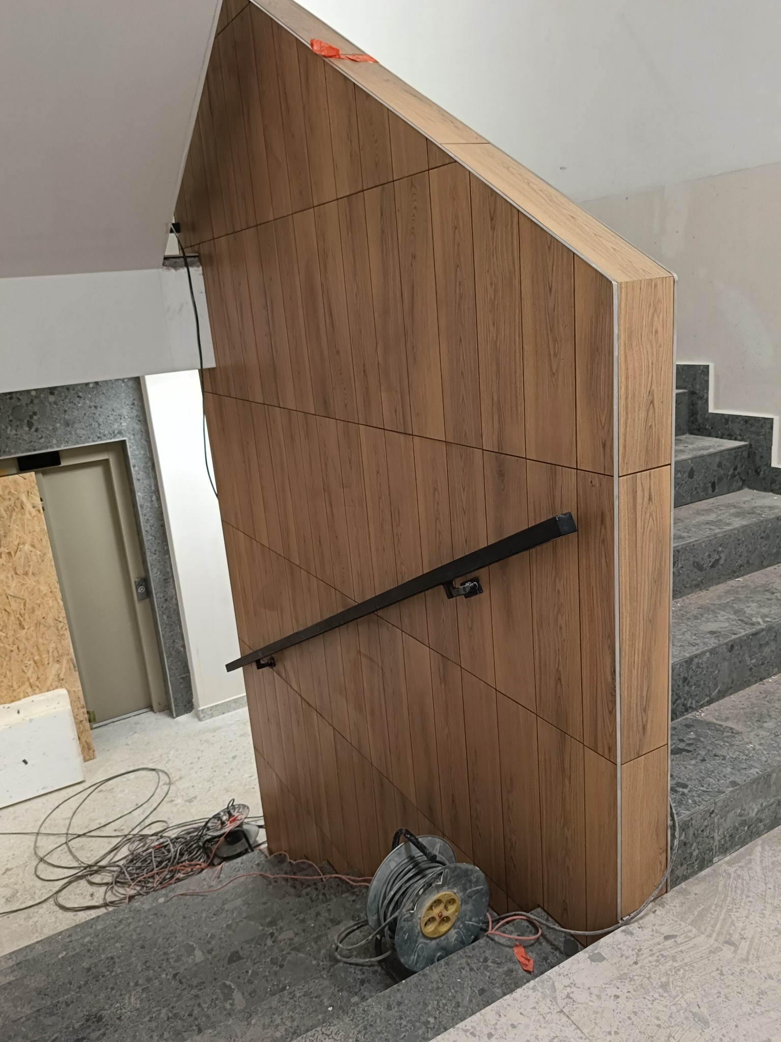 Wykończenie klatki schodowej płytkami drewnopodobnymi na ścianie i schodach z szarego lastryko, czarna poręcz, zwinięty przedłużacz na schodach, widoczne kable elektryczne.