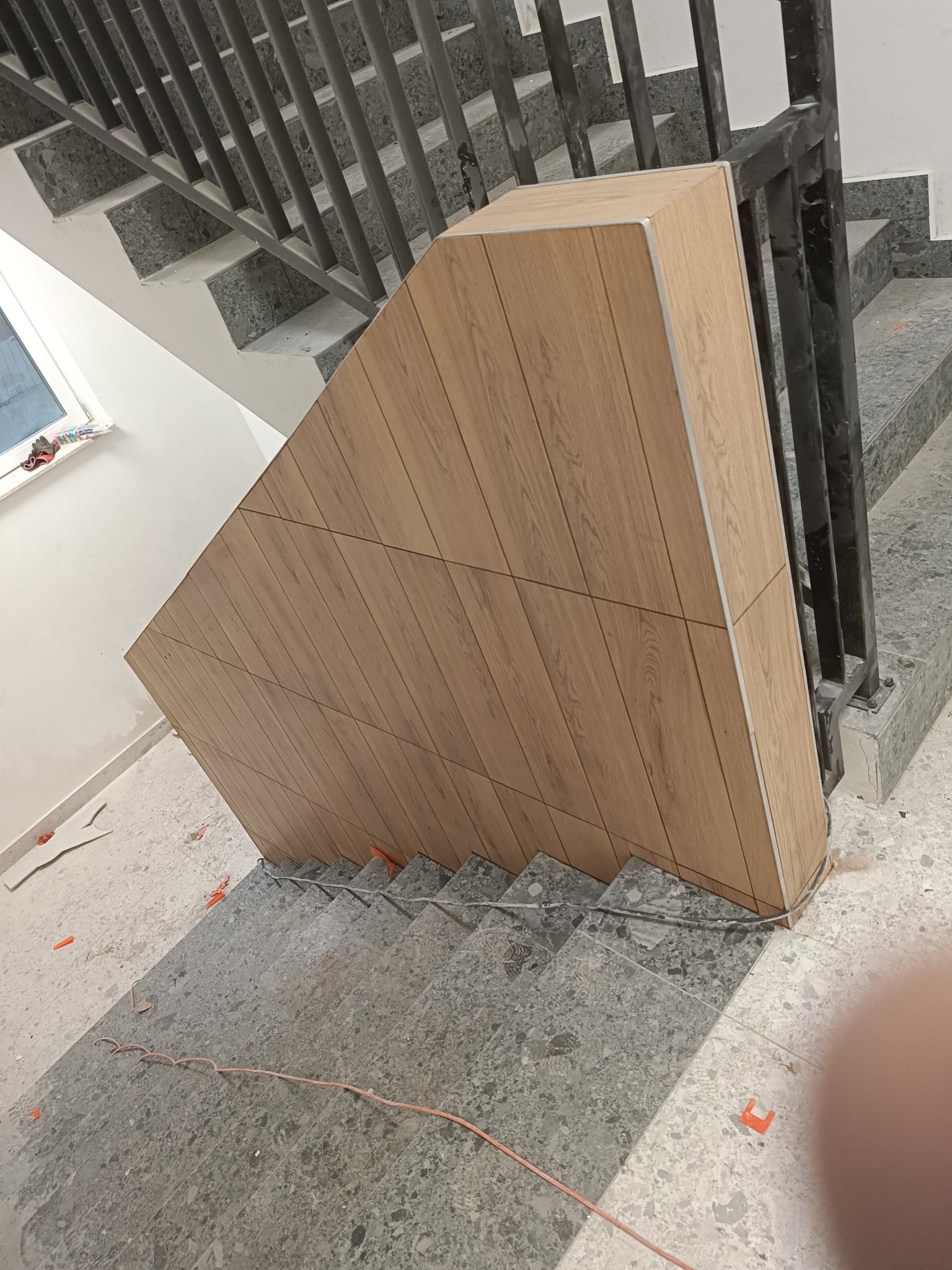 Wykończenie klatki schodowej panelami imitującymi drewno, widoczne szare schody z lastryko i czarna metalowa balustrada, w tle okno i biała ściana, na schodach widoczne elementy montażowe.