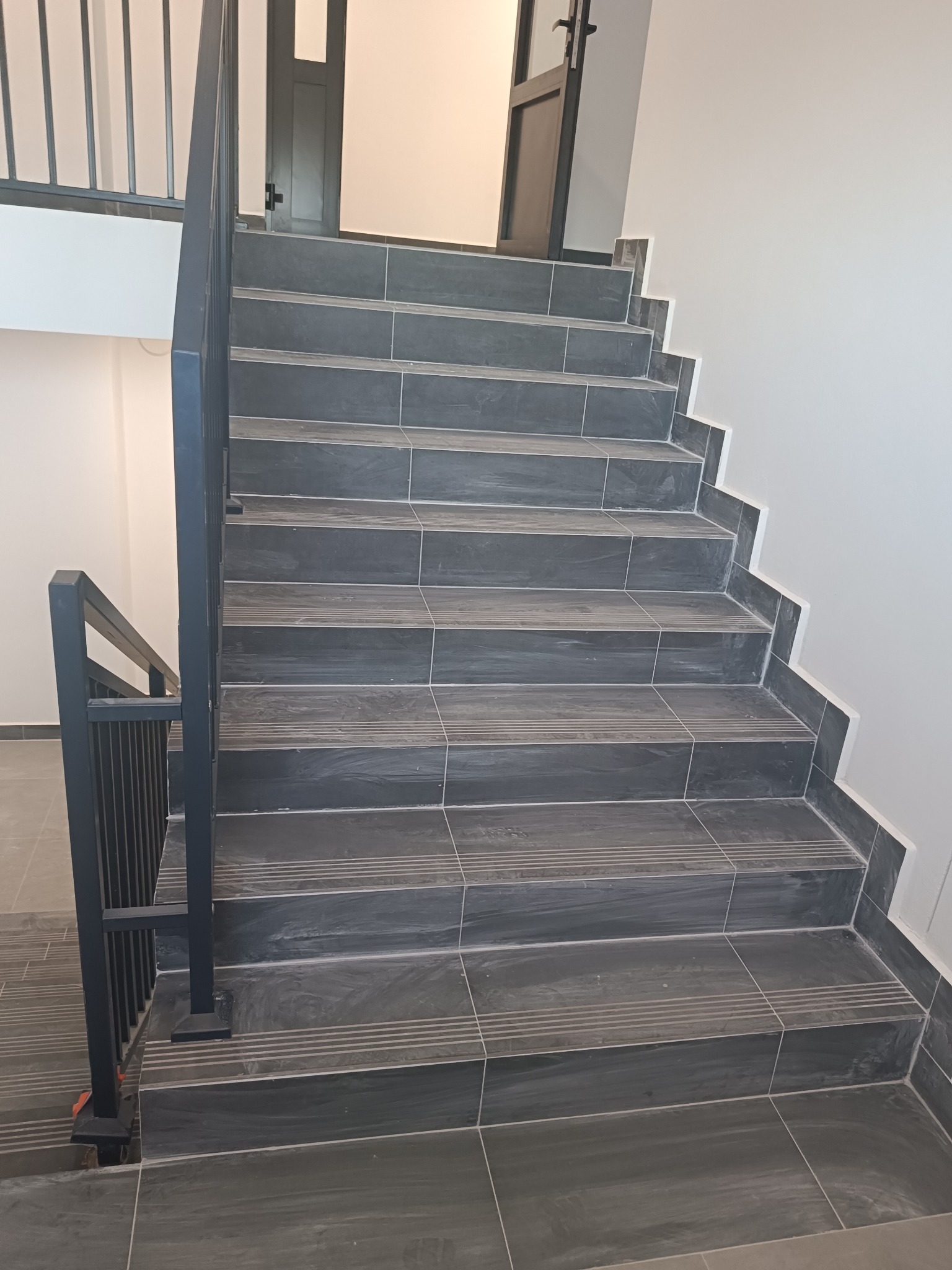 Schody wyłożone ciemnymi płytkami z antypoślizgowymi rowkami, czarna metalowa balustrada, widok z dołu na górę, białe ściany.