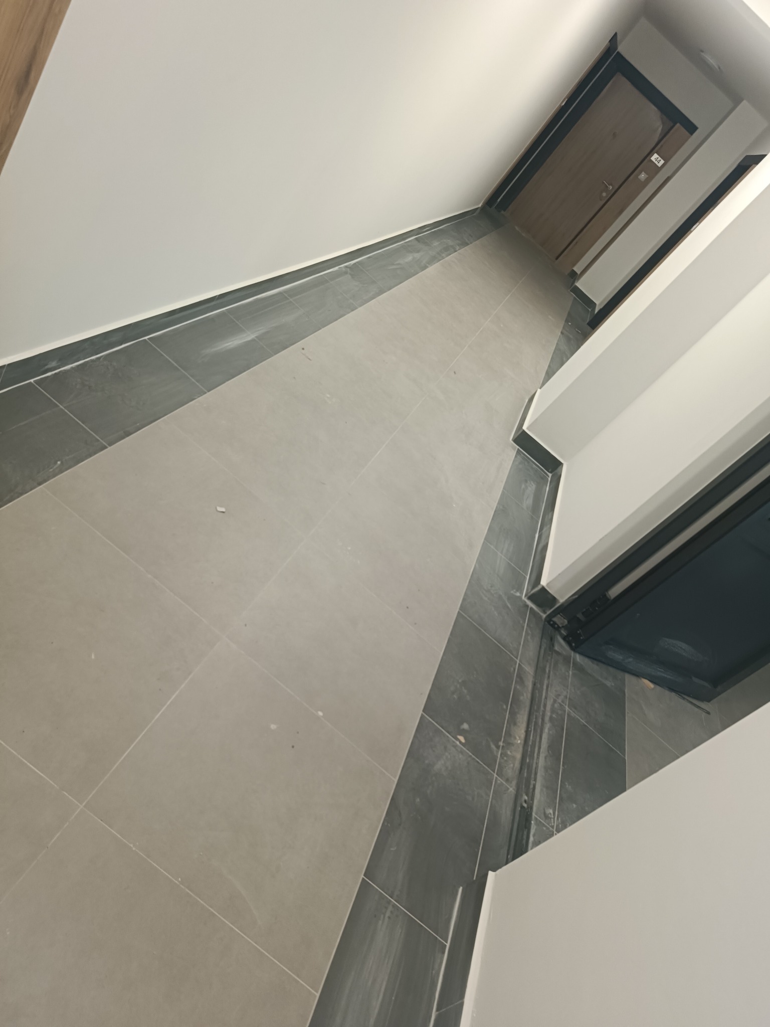 Korytarz wyłożony jasnymi, kwadratowymi płytkami z ciemnoszarym obramowaniem, prowadzący do drzwi z numerem '44'.