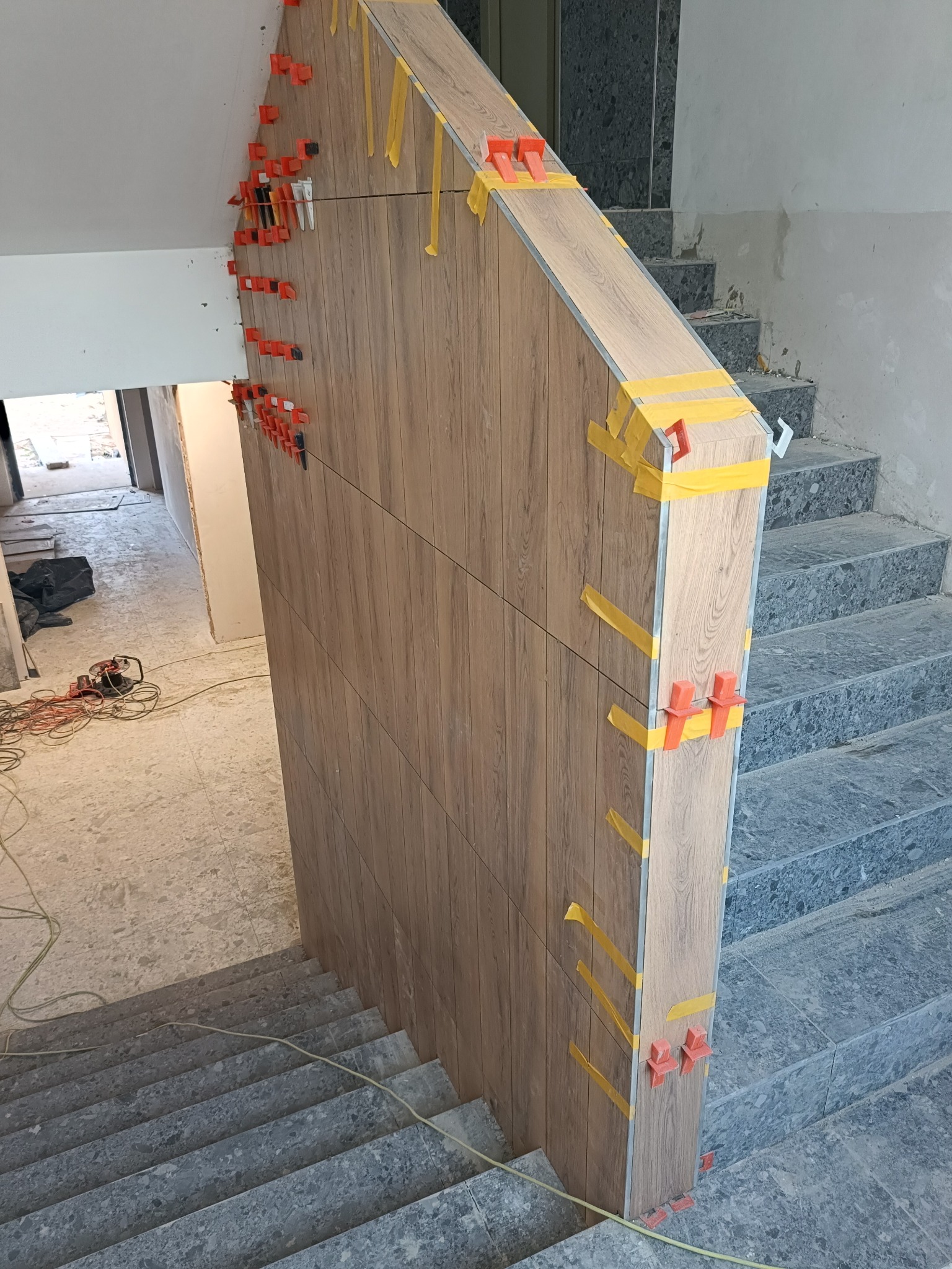 Montaż drewnopodobnych płytek na schodach z użyciem klipsów poziomujących i taśmy maskującej, widoczne narzędzia w tle, etap prac wykończeniowych.