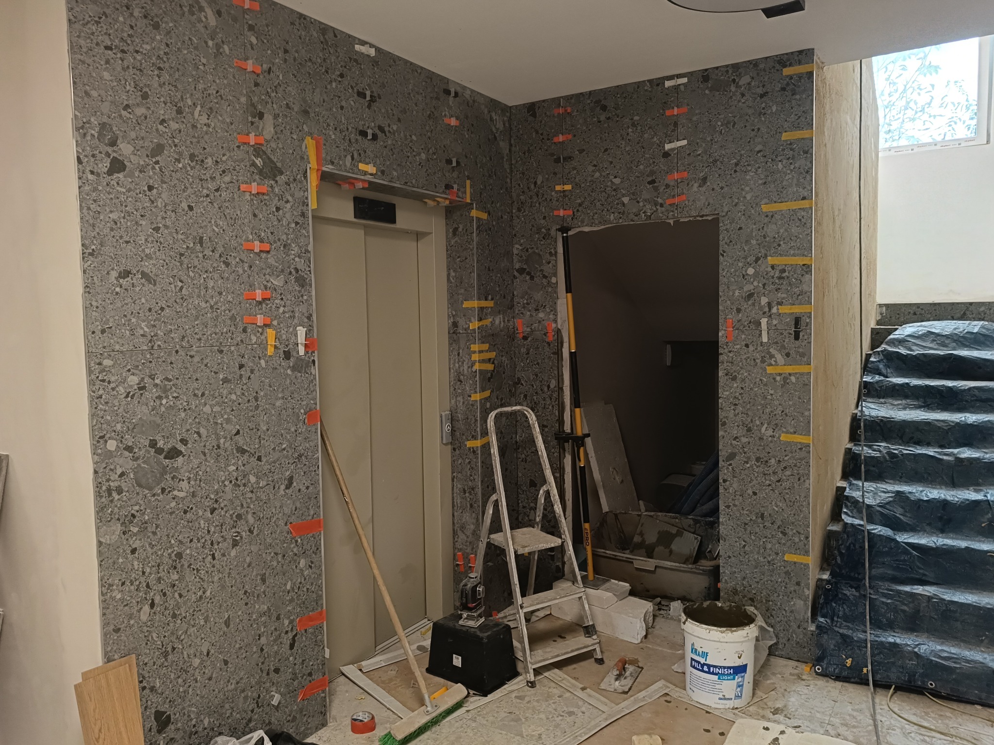 Montaż szarej mozaiki na ścianie przy windzie i schodach, z widocznymi plastikowymi krzyżykami dystansowymi, drabina, wiadro z masą szpachlową i narzędzia glazurnicze na podłodze.