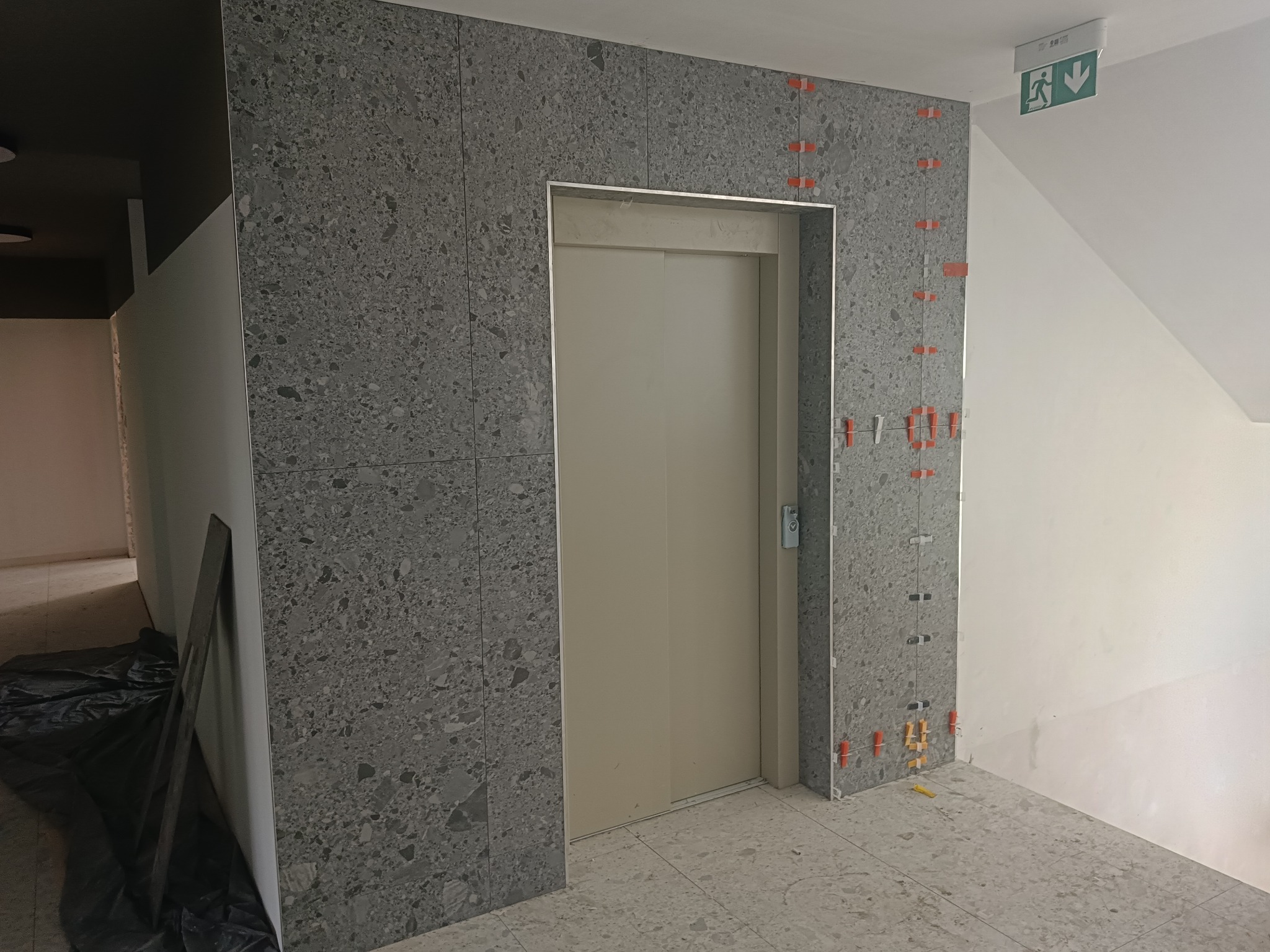 Montaż płytek z efektem lastryko wokół windy, z użyciem krzyżyków dystansowych, widoczne płyty do dalszego montażu i znak ewakuacyjny.