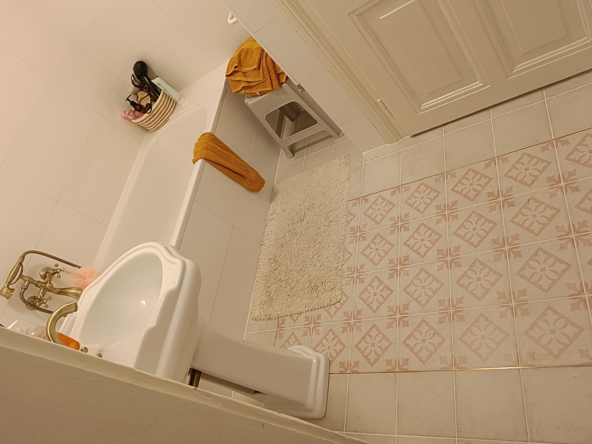 Ukośne ujęcie łazienki z umywalką na postumencie, złotą armaturą, beżowymi płytkami podłogowymi w kwiatowy wzór i akcesoriami w koszyku nad wanną.