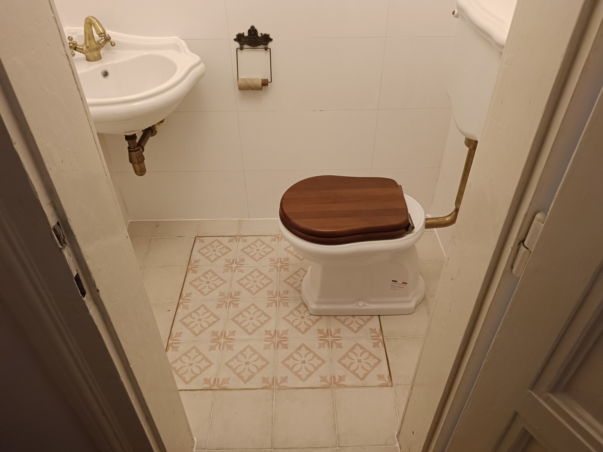 Retro łazienka z białą ceramiką, drewnianą deską sedesową, umywalką z mosiężną armaturą i wzorzystymi płytkami podłogowymi, widok zza uchylonych drzwi.