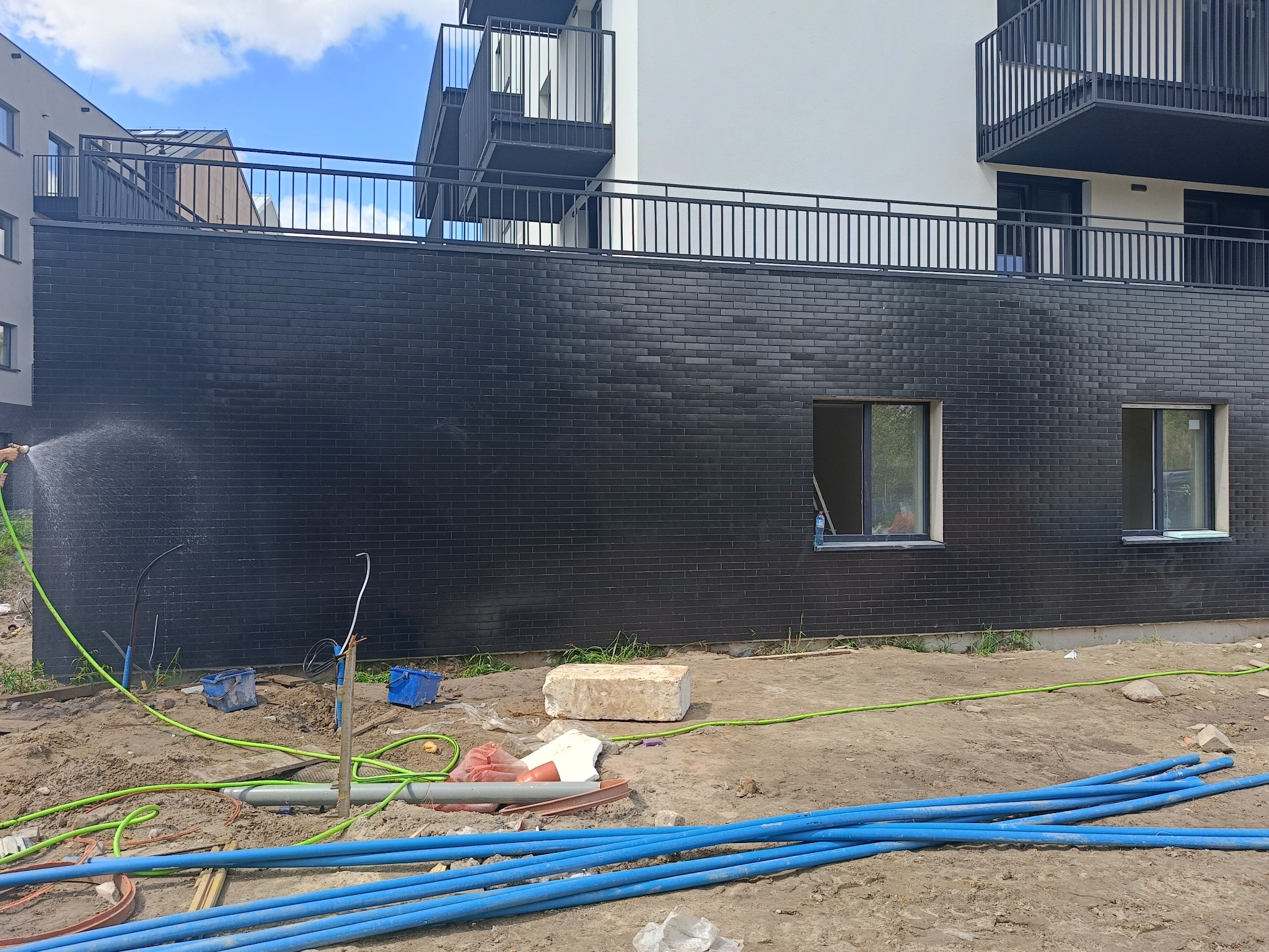 Czyszczenie elewacji budynku z czarnej cegły klinkierowej po zakończeniu prac, z widocznymi oknami i balkonami, oraz narzędziami i materiałami budowlanymi na ziemi.