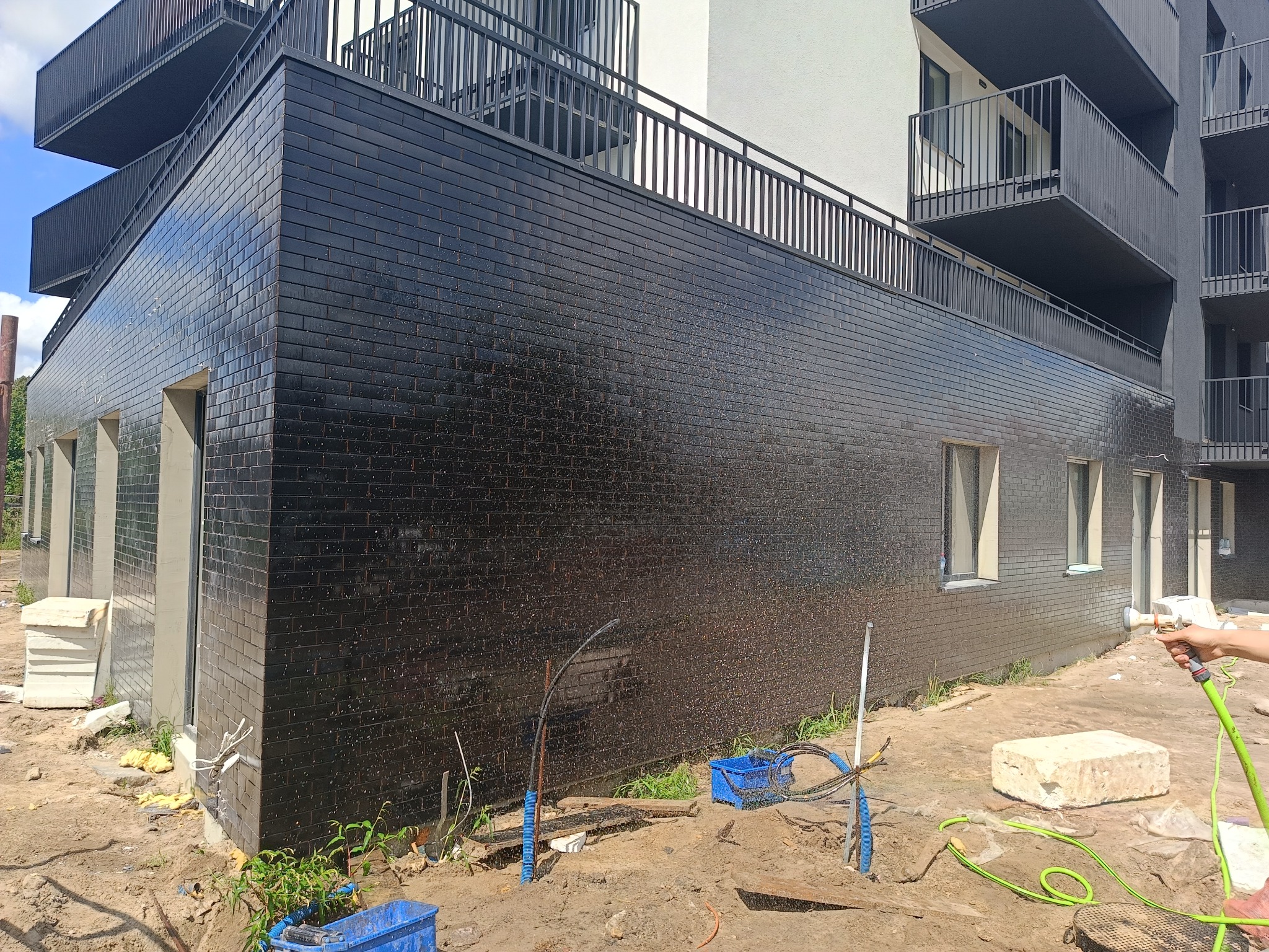 Nowoczesny budynek wielorodzinny z czarną, błyszczącą elewacją z cegły klinkierowej, myty wodą pod ciśnieniem po zakończeniu prac, widoczne elementy instalacji i narzędzia na placu budowy.