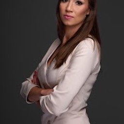 Kancelaria Radcy Prawnego Justyna Rembisz - Portret kobiety w jasnej sukience z rękami skrzyżowanymi na piersiach, stojącej na ciemnoszarym tle.