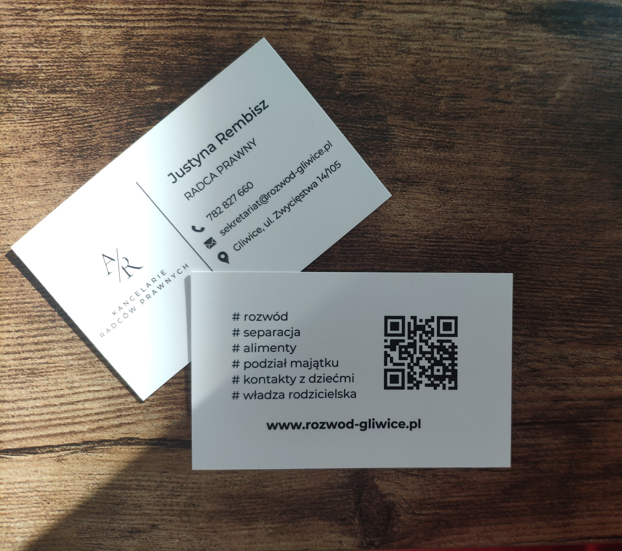 Dwie wizytówki radcy prawnego Justyny Rembisz na drewnianym tle, oferującej usługi związane z rozwodem, separacją, alimentami, podziałem majątku, kontaktami z dziećmi i władzą rodzicielską,...