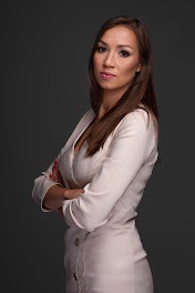 Portret kobiety w jasnej sukience z rękami skrzyżowanymi na piersiach, stojącej na ciemnoszarym tle.