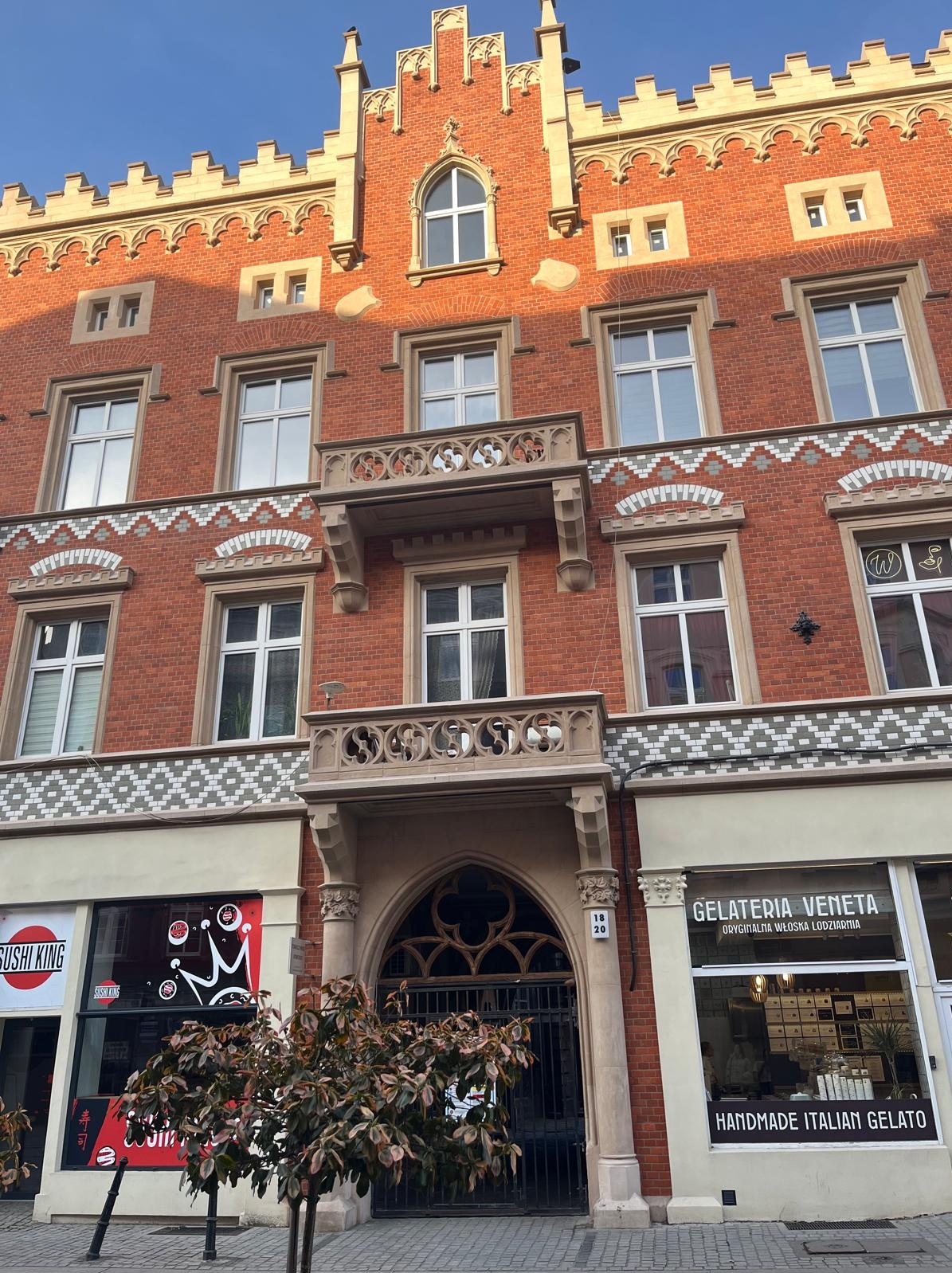 Kamienica z czerwonej cegły z dekoracyjnymi balkonami i gotyckim portalem wejściowym, poniżej lokale usługowe Sushi King i Gelateria Veneta.