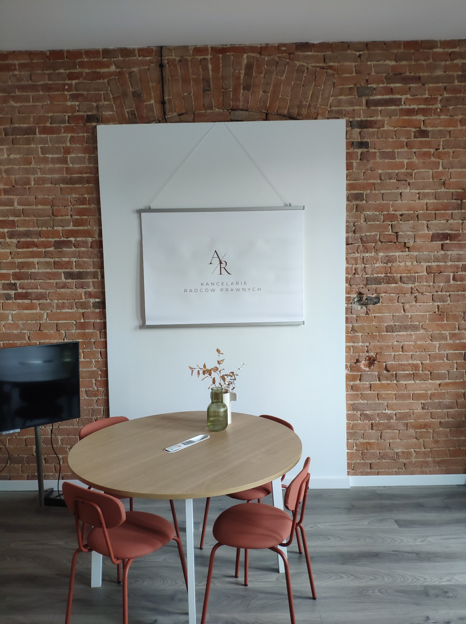 Wnętrze kancelarii z ceglaną ścianą, okrągłym stołem i plakatem z logo kancelarii radców prawnych.
