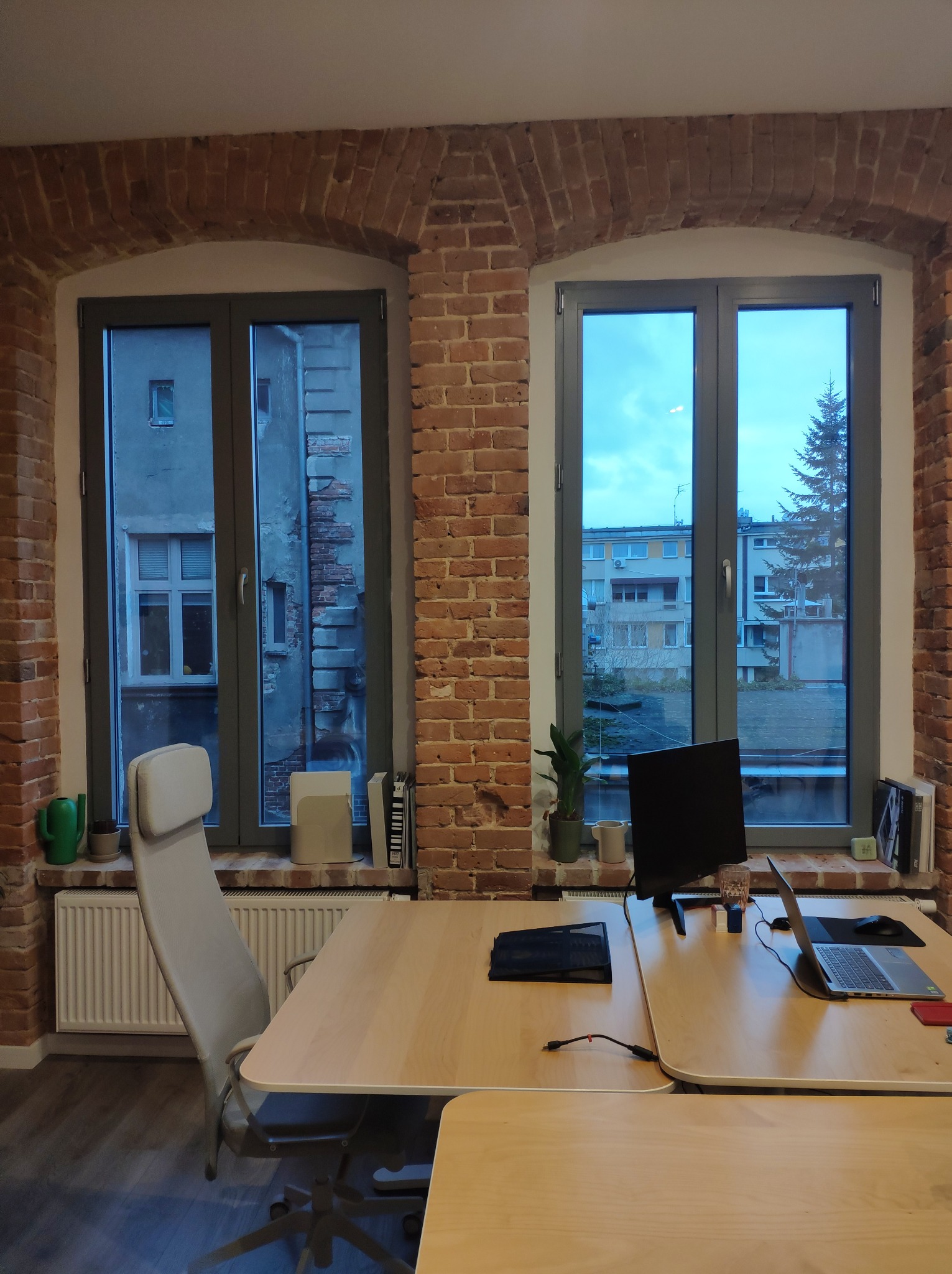Biuro z ceglanymi ścianami, dwoma oknami z widokiem na budynki i drzewo, biurkiem z laptopem i monitorem oraz ergonomicznym krzesłem.