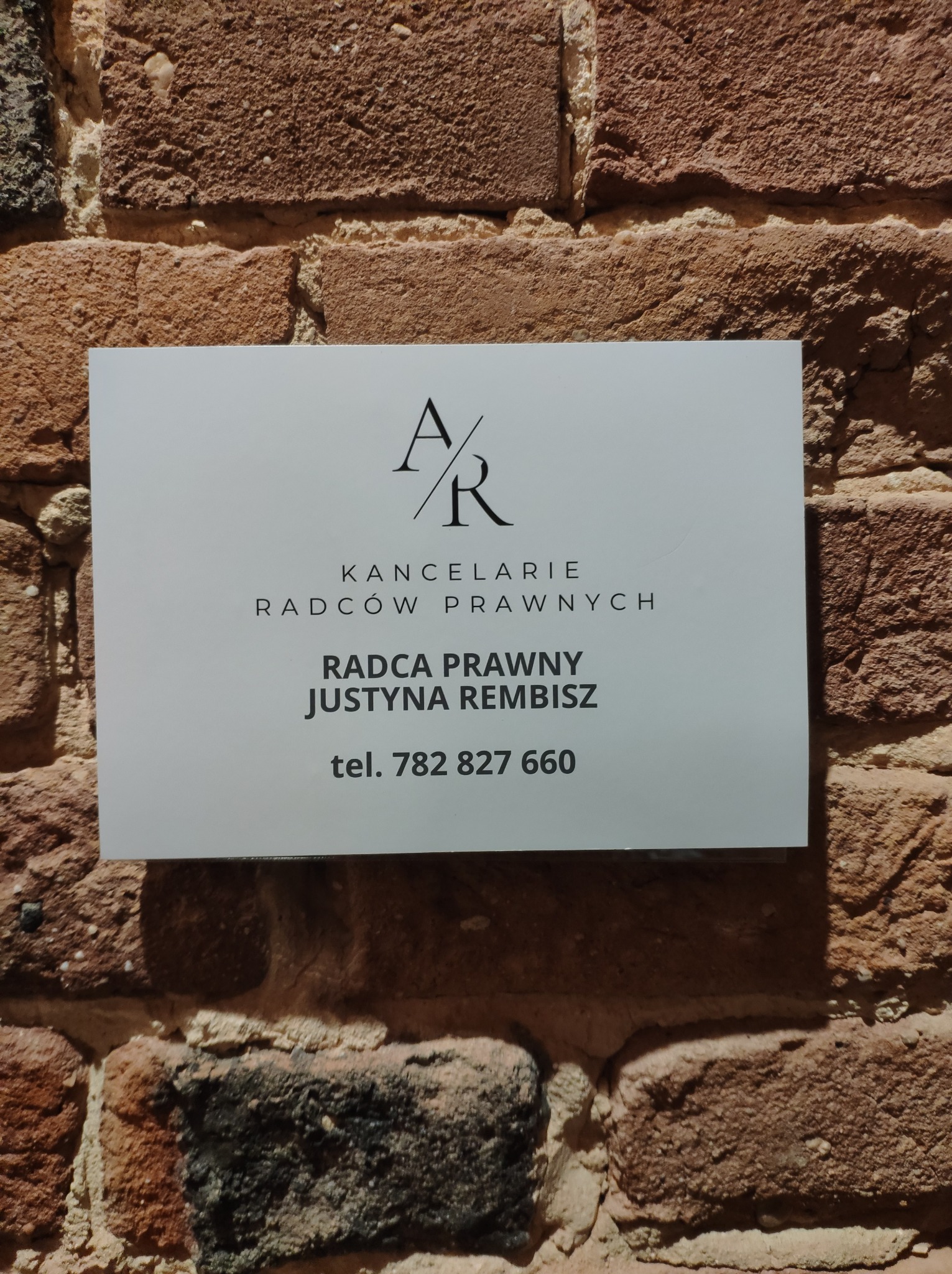 Tabliczka informacyjna Kancelarii Radców Prawnych Justyna Rembisz, umieszczona na ścianie z czerwonej cegły, z widocznym logo AR i numerem telefonu.