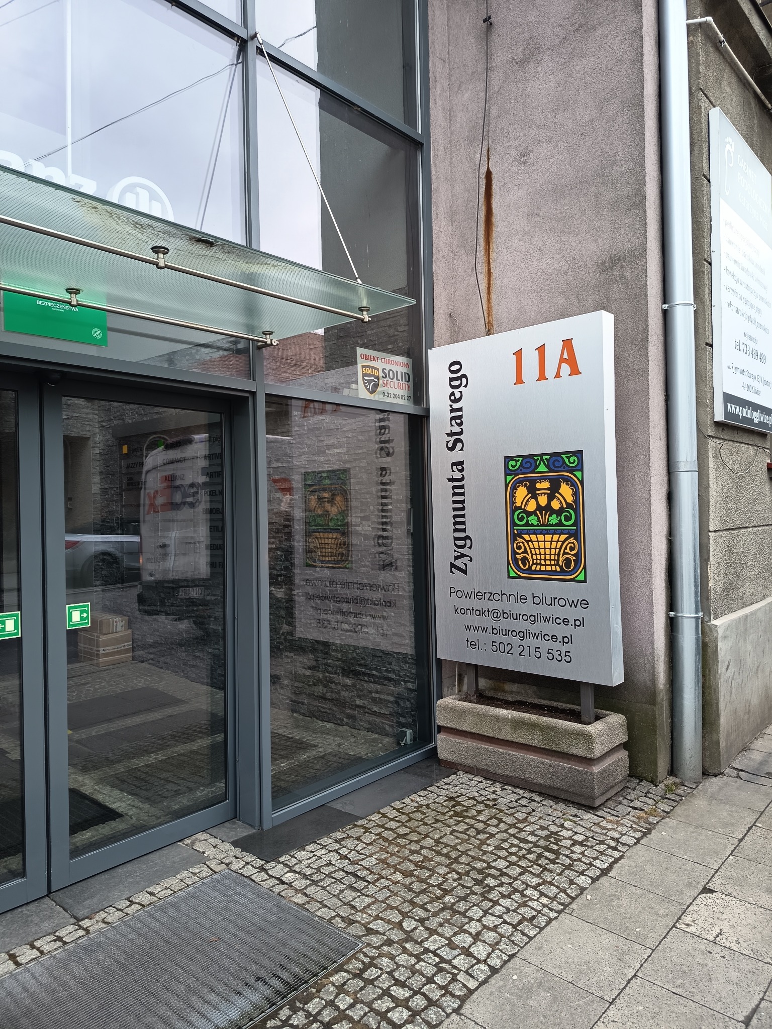 Szyld z adresem 'Zygmunta Starego 11A' przed wejściem do biura, z ozdobnym motywem roślinnym i danymi kontaktowymi. Widoczny fragment chodnika z kostki brukowej.
