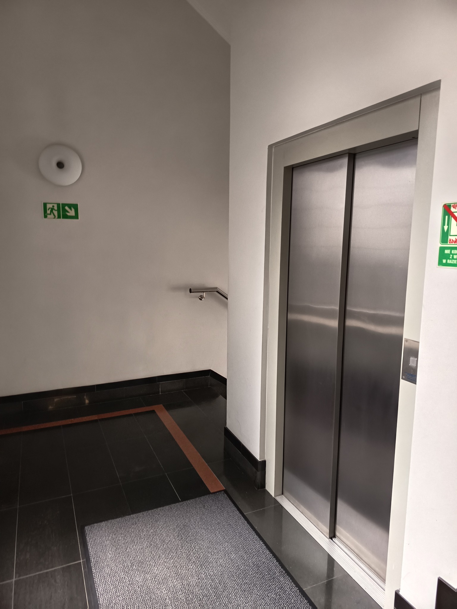 Wnętrze budynku z windą o metalowych drzwiach, czarną podłogą i szarą wycieraczką. Na ścianie znak ewakuacyjny i lampa.