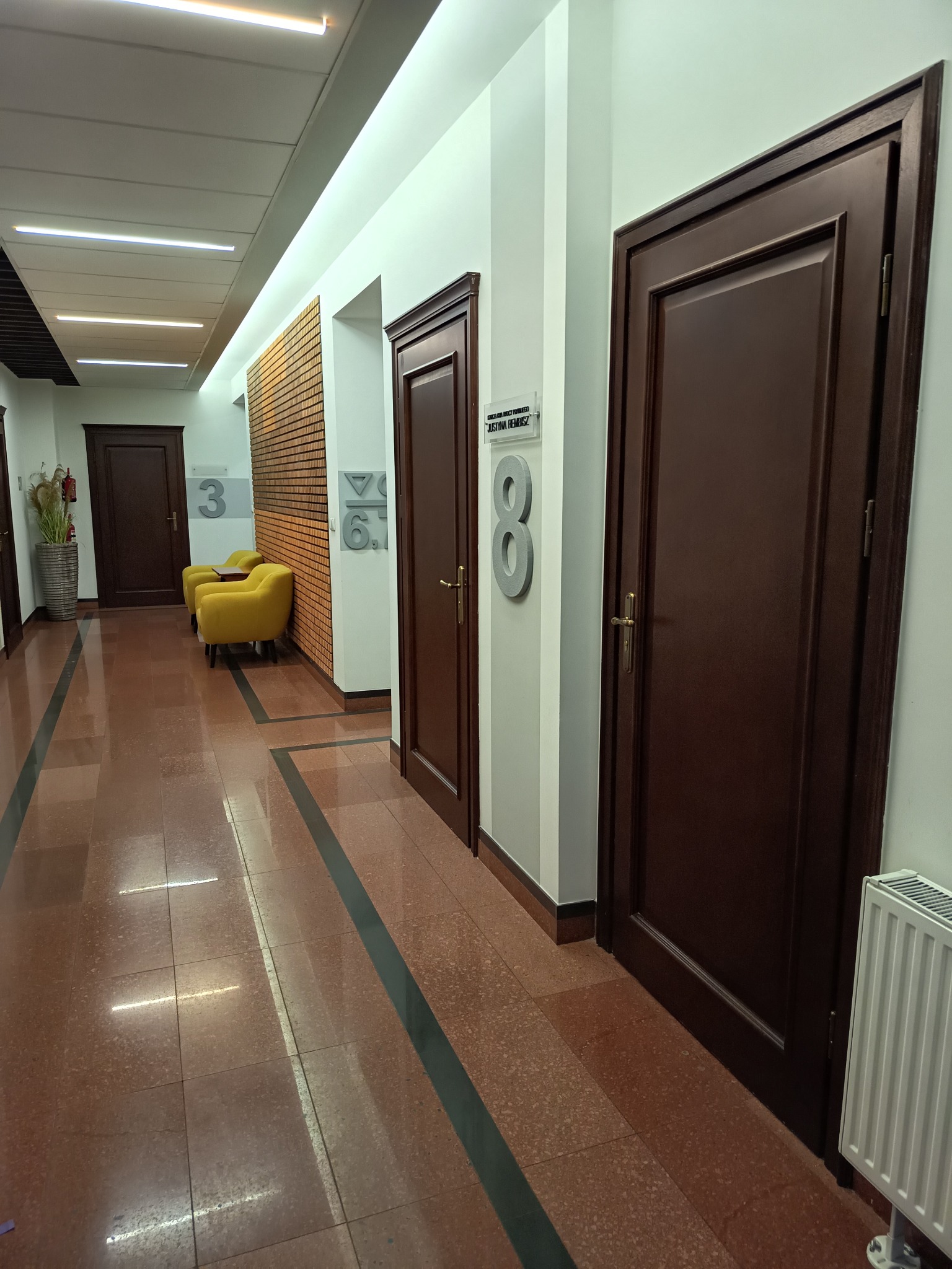 Korytarz kancelarii prawnej z numerami gabinetów, żółtymi fotelami i brązowymi drzwiami. Widoczny fragment grzejnika i podłoga z terakoty.