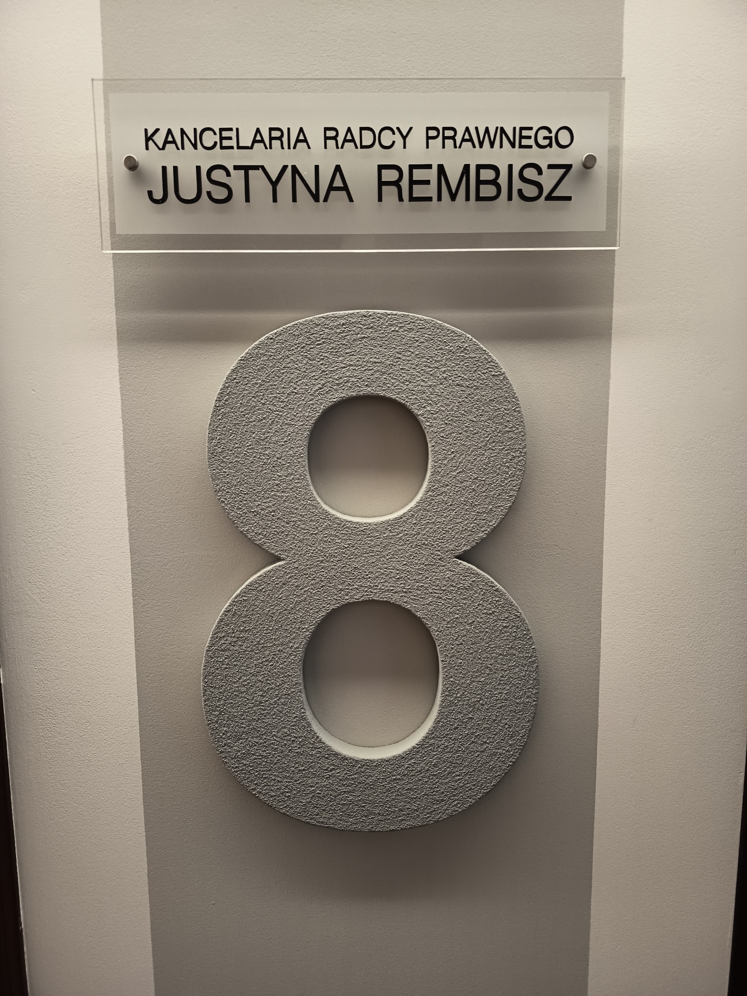 Tabliczka z napisem 'Kancelaria Radcy Prawnego Justyna Rembisz' nad numerem '8' na ścianie. Minimalistyczny wystrój, stonowana kolorystyka.