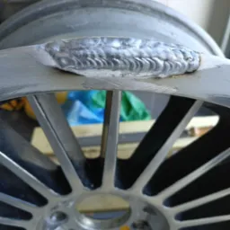 Szlif spawalniczy na obręczy aluminiowej. Widoczne ślady spawania i obróbki metalu. Obręcz z wieloma szprychami.