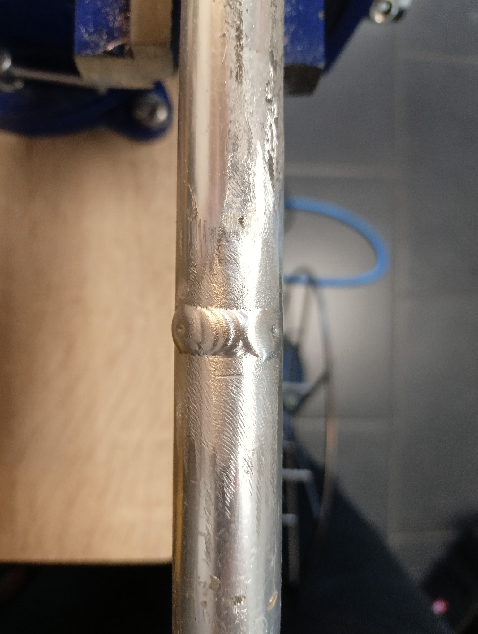 Spoina na metalowej rurze uchwyconej w imadle. Widoczne ślady obróbki i niedoskonałości spoiny. W tle elementy warsztatu.